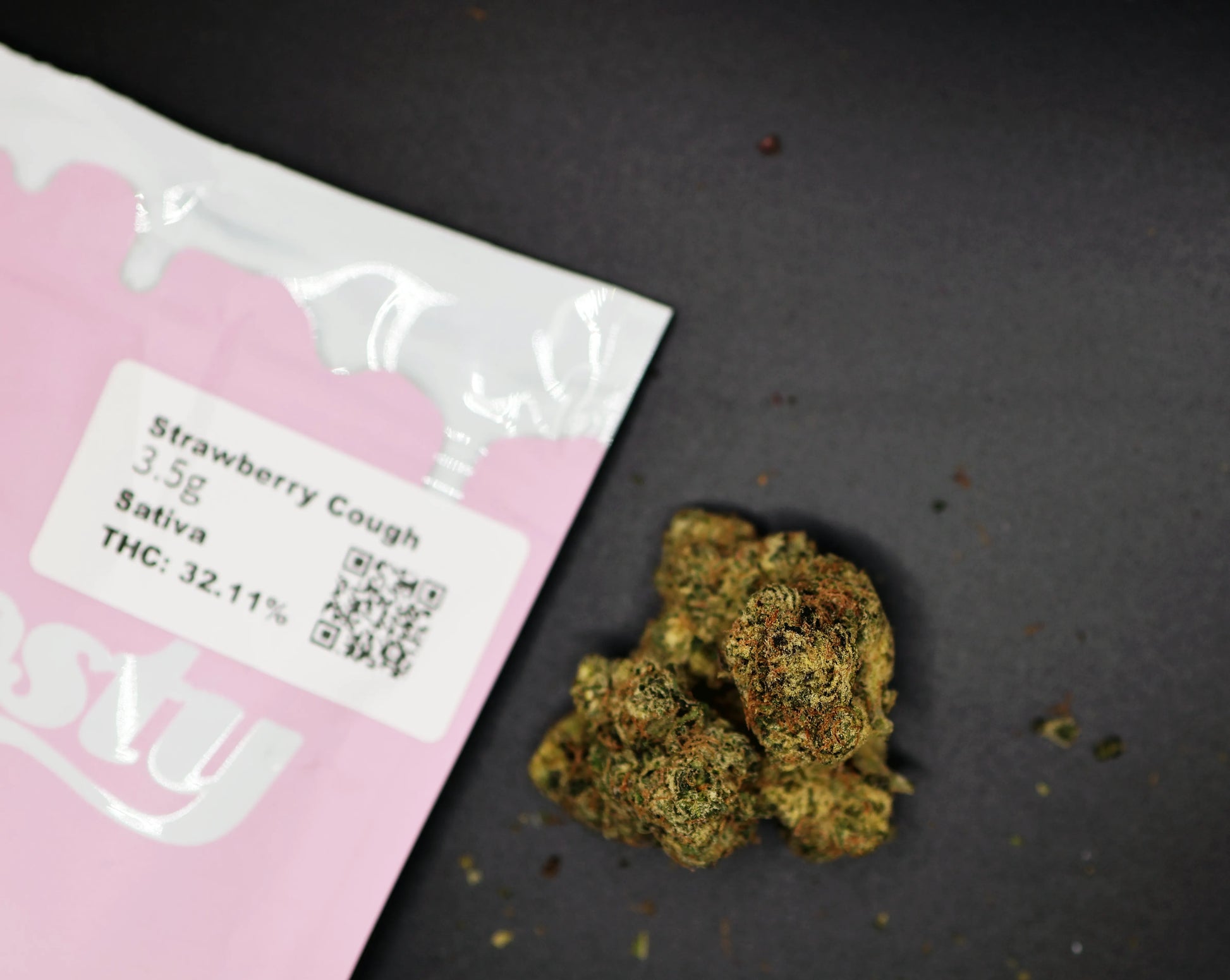Pink packaging of 'Strawberry Cough' Sativa with cannabis buds on a dark surface