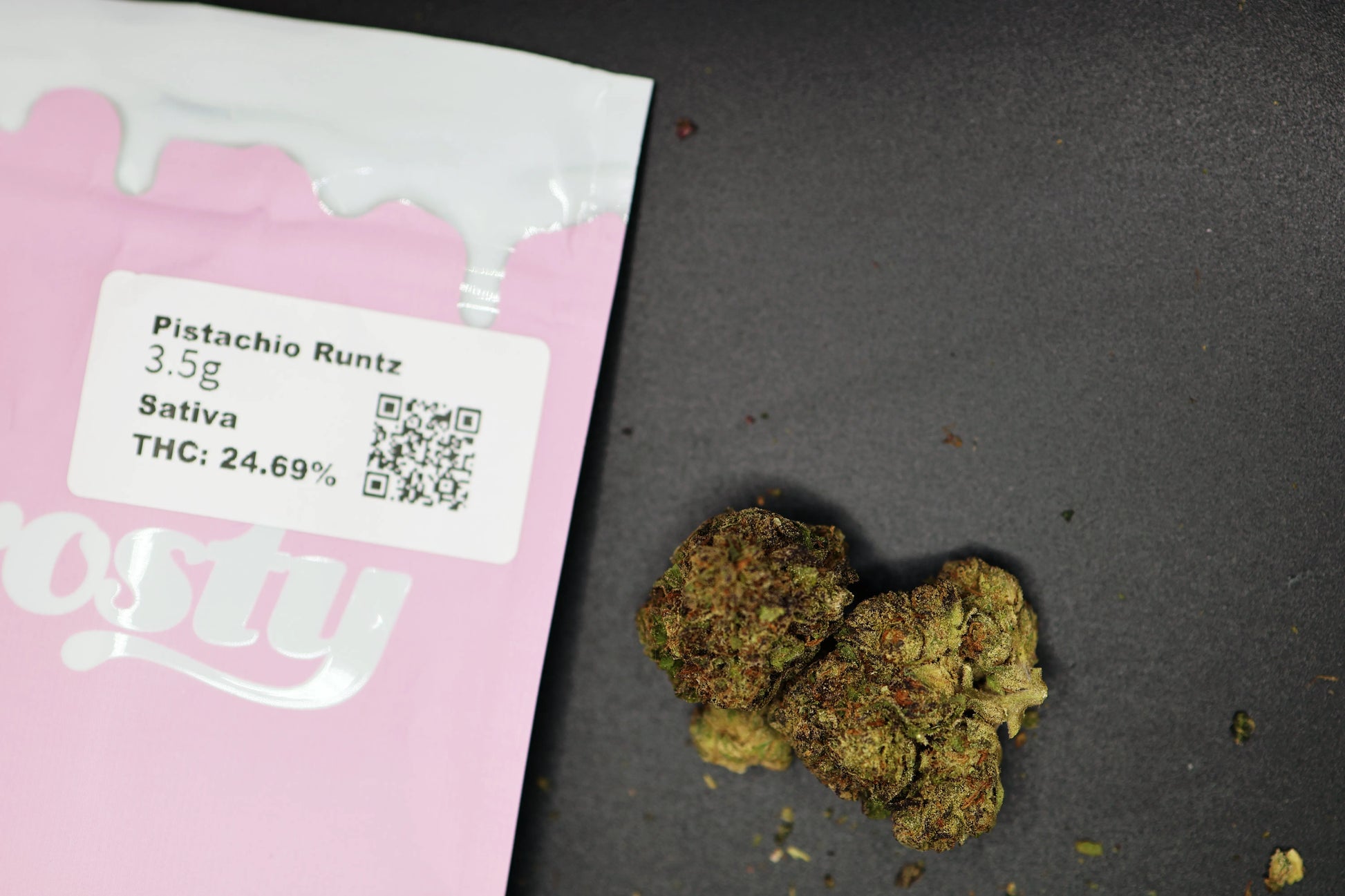 Marijuana buds next to a pink packaging with product details on a dark surface, pistachio runtz strain